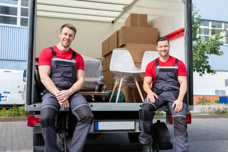 Portrait Of Smiling Movers Sitting In Van With Stack Of Cardboard Boxes And Furniture
