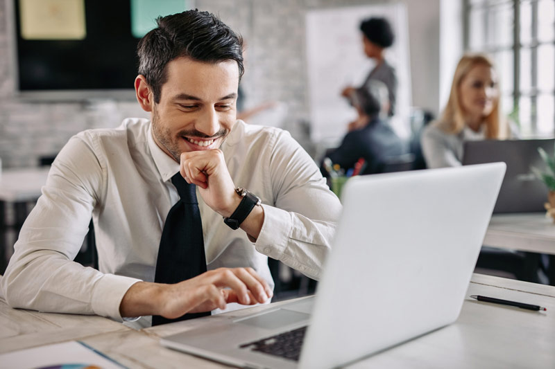 web smiling businessman sitting desk office working laptop there are people background 1